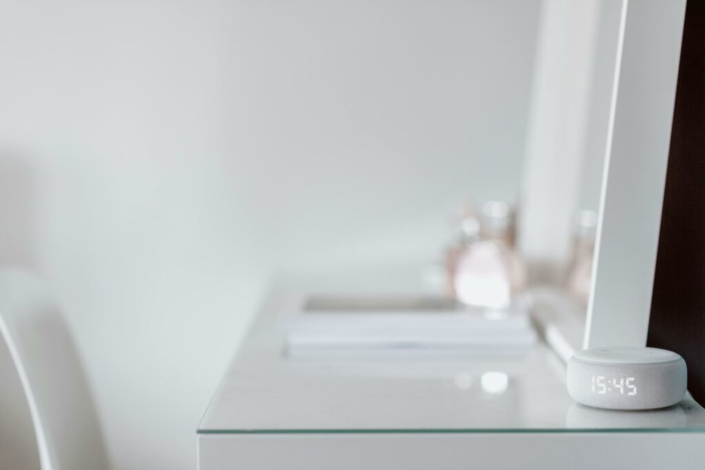 Smart speaker with LED clock on a white desk, next to a mirror and perfume bottles in a modern smart home setting.