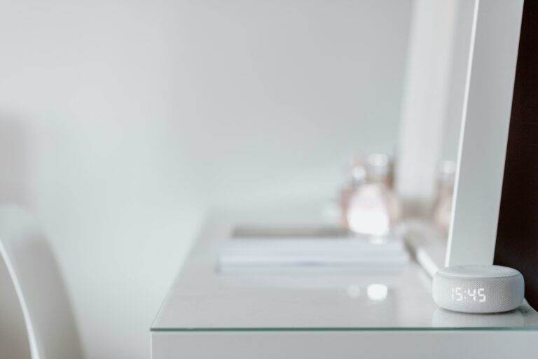Smart speaker with LED clock on a white desk, next to a mirror and perfume bottles in a modern smart home setting.